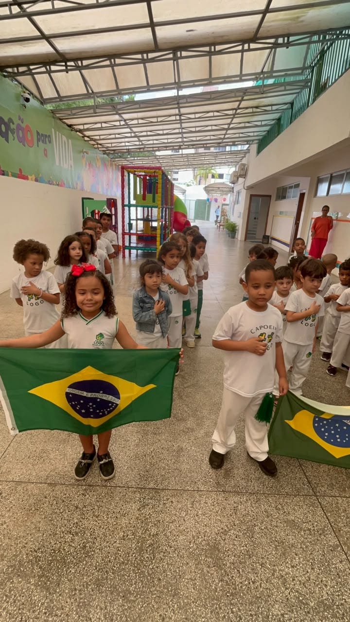 Fila de crianças no corredor, de uniforme branco de capoeira, com a bandeira do Brasil