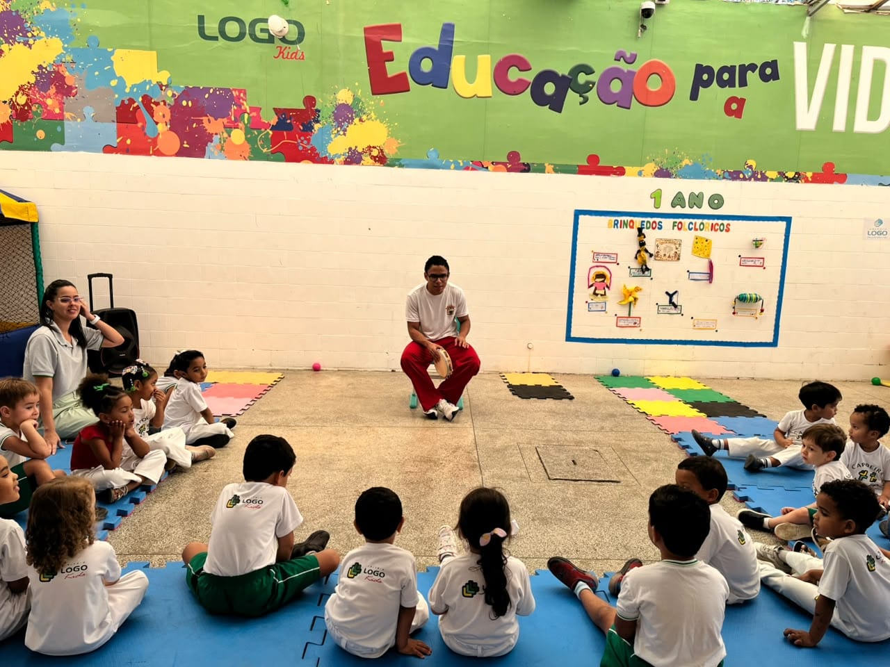 Roda com o professor diante do painel de brinquedos folclóricos, crianças sentadas no chãoa à frente