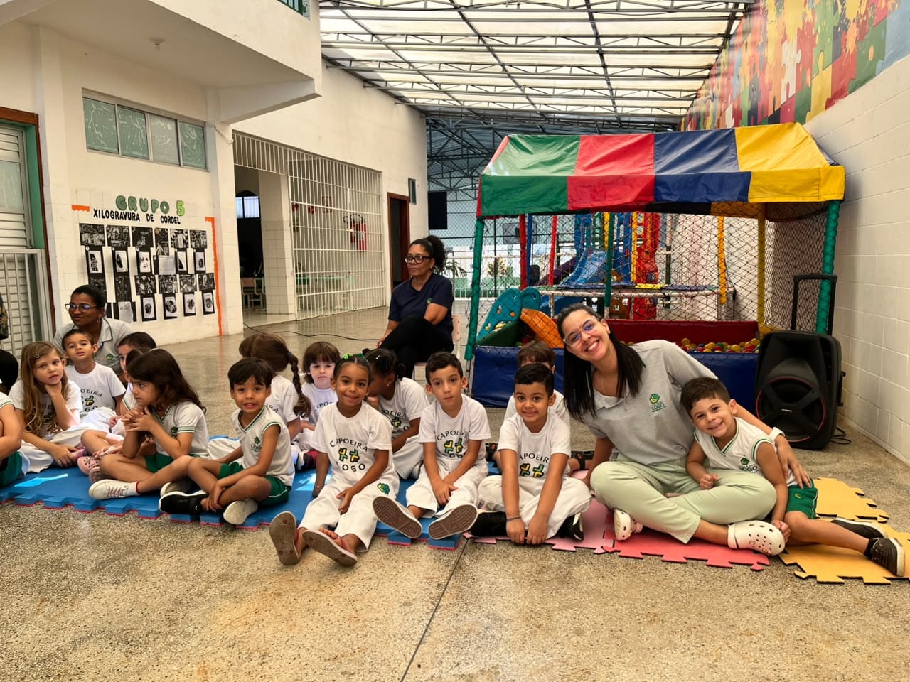 Crianças de uniforme de capoeira sentadas nos tatames coloridos da área coberta, com a professora