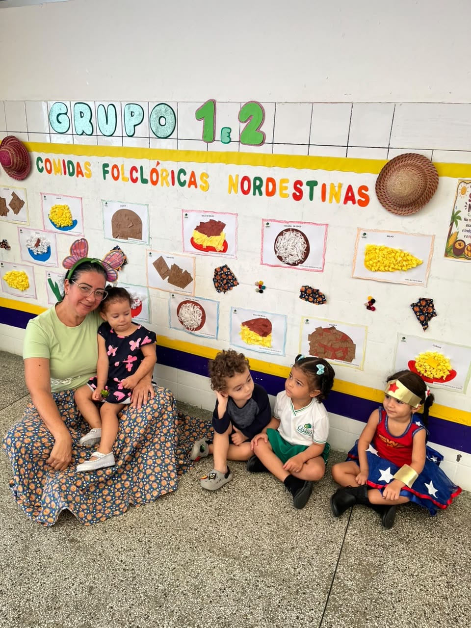Professora sentada com uma criança no colo, diante do painel de comidas folclóricas