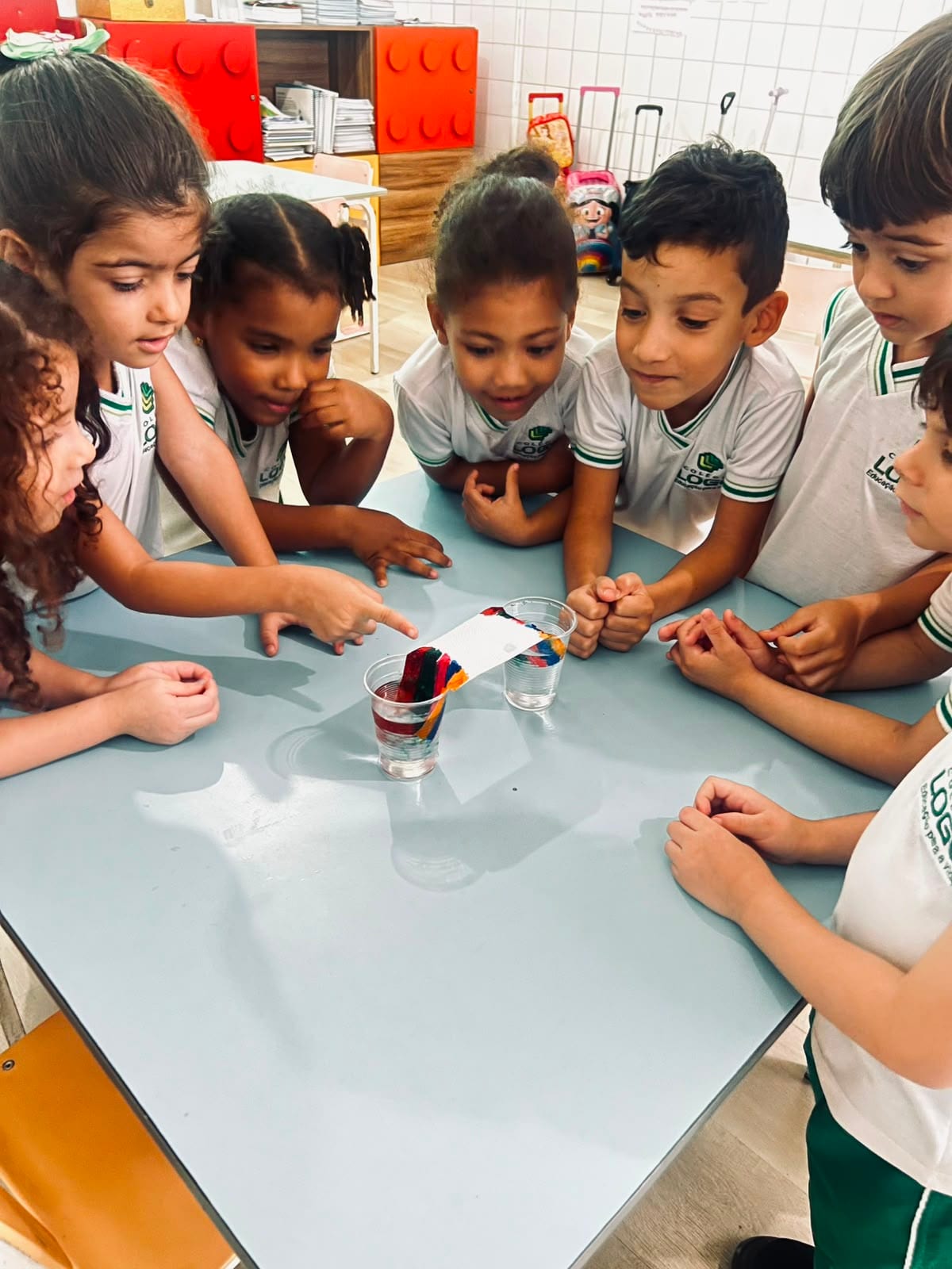 Crianças em volta de uma mesa observando um copo com líquidos coloridos em camadas