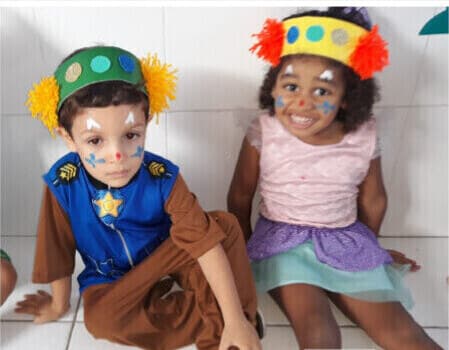 Menino fantasiado de policial e menina de vestido rosa sentados no chão, com adereços coloridos na cabeça